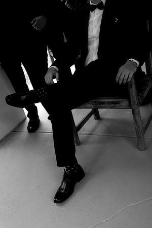 Black and white photo of a man in a tuxedo and bow tie sitting on a wooden chair, adjusting his dress shoes.