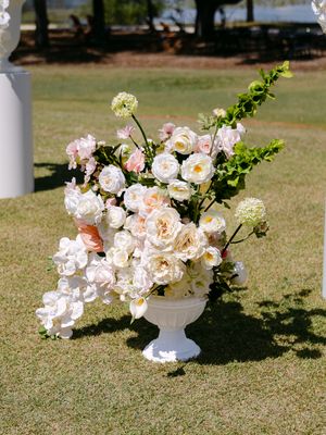 White urn vase with lush blush pink and white roses, hydrangeas, and greenery arrangement on grass at outdoor wedding.