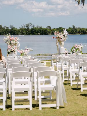 Outdoor wedding ceremony setup with white folding chairs, floral arrangements, and a waterfront backdrop under a blue sky.