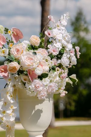 Elegant white urn vase with pink and white roses, orchids, and mixed blooms outdoors at a wedding ceremony.