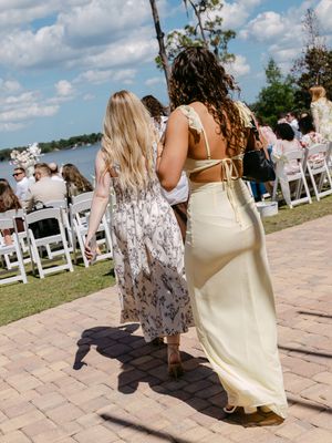 Two women in elegant dresses walk toward an outdoor wedding ceremony by a lakeside on a sunny day.