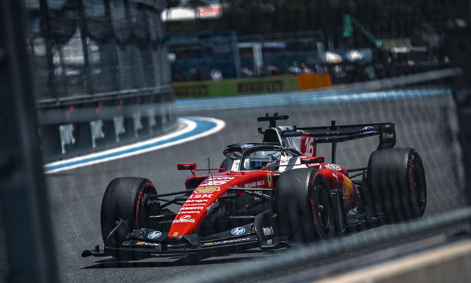Red Ferrari F1 car racing on a wet track, captured through a fence at a Grand Prix circuit.