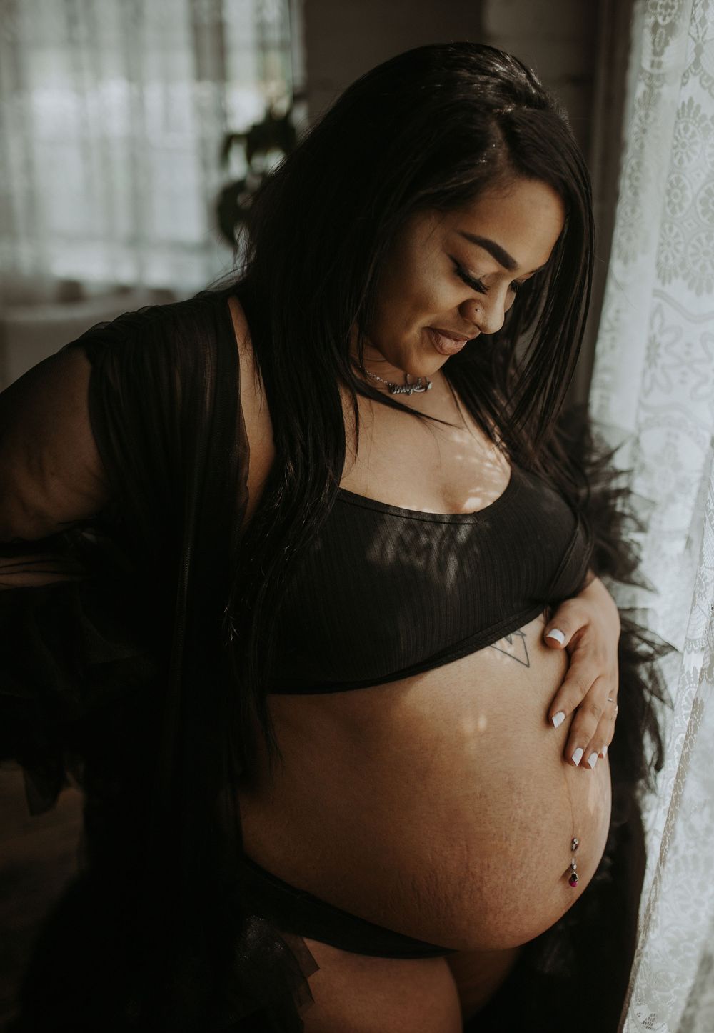 Pregnant woman in black lingerie and sheer robe smiling while cradling baby bump by window.