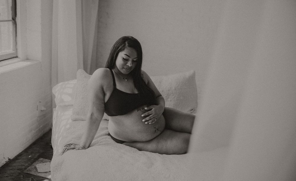 Black and white maternity photo of a pregnant woman sitting on a bed, cradling her baby bump.