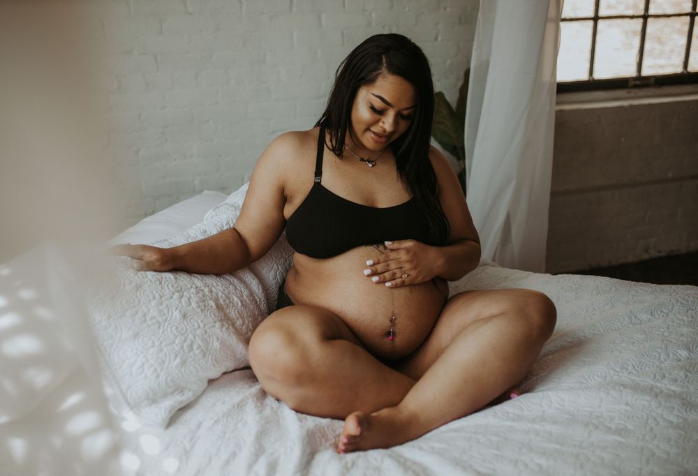 Pregnant woman in black bralette sitting cross-legged on white bed cradling her baby bump.