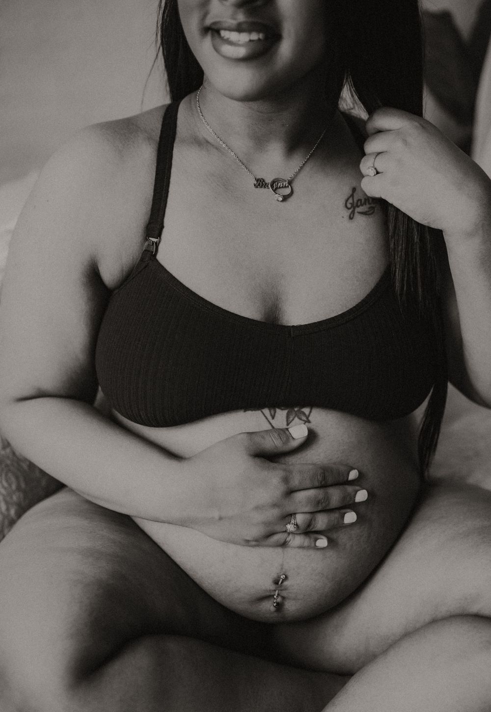 Black and white photo of pregnant woman in black bralette holding her belly with manicured nails.