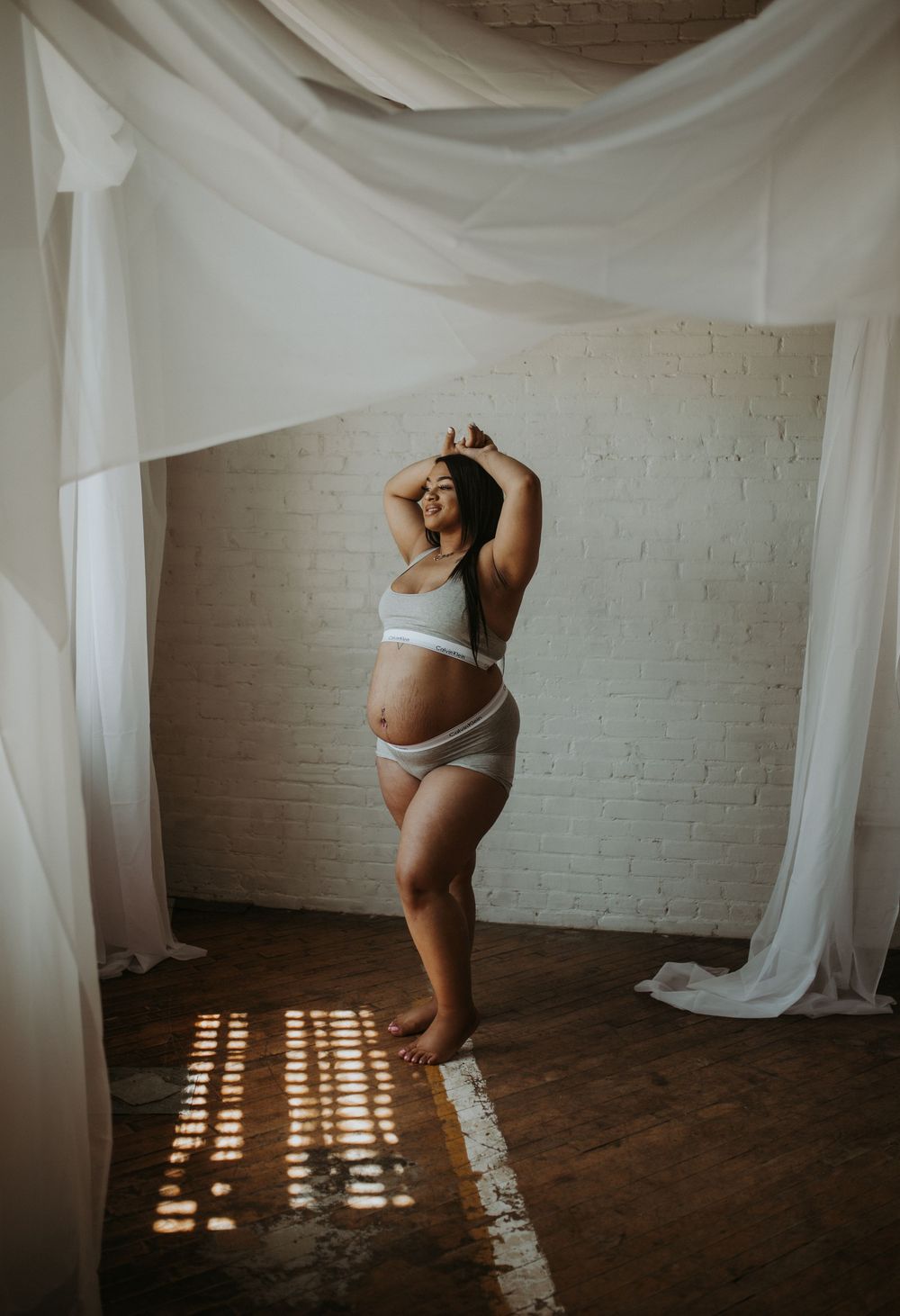 Smiling pregnant woman in white lingerie poses under flowing white fabric drapes.