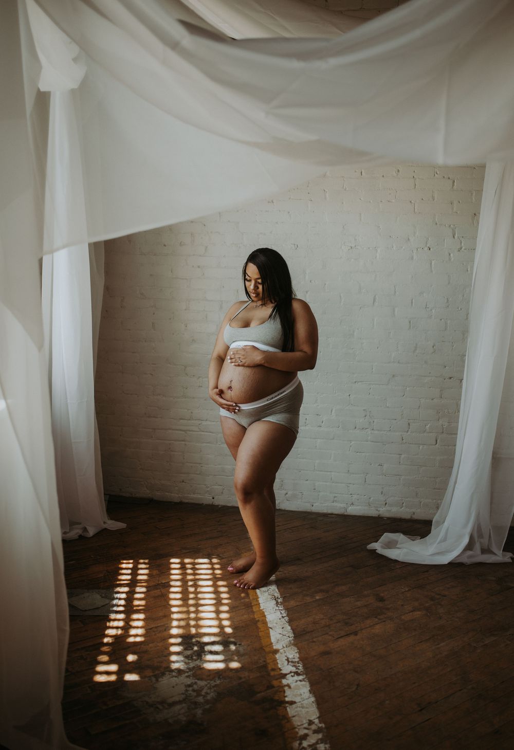 Pregnant woman in white lingerie cradling baby bump, standing in sunlit studio with white drapes and brick wall.