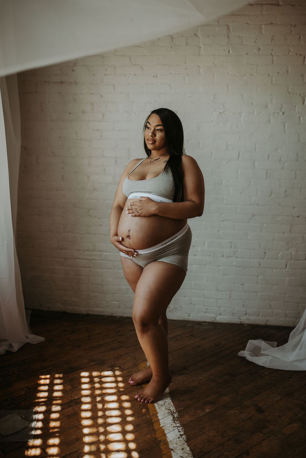 Pregnant woman in white lingerie standing against white brick wall in sunlight.