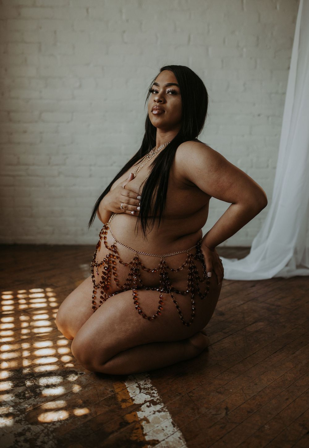 Pregnant woman kneeling on wood floor wearing decorative body chain jewelry in warm sunlight.