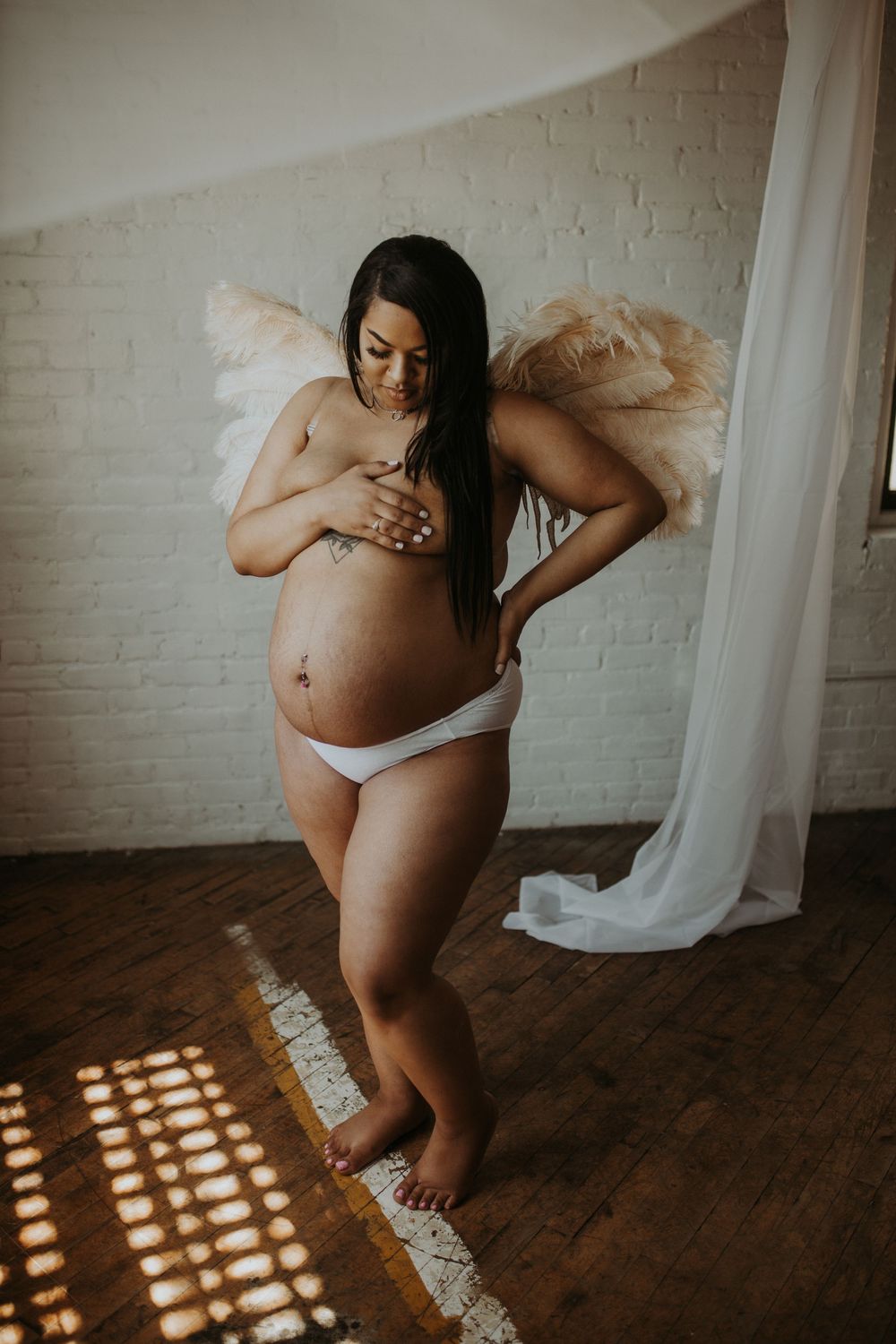 Pregnant woman wearing angel wings and white underwear cradling her bare baby bump indoors.