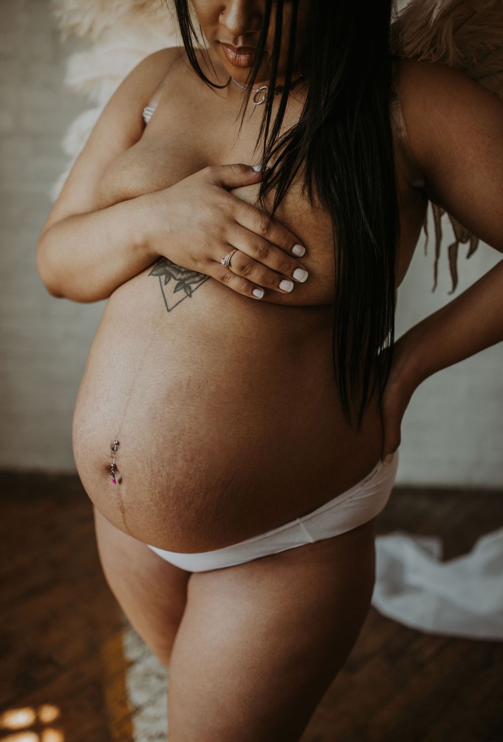 Close-up of pregnant belly with tattoo and belly piercing, woman covering chest.