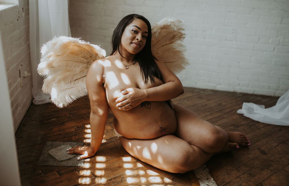 Pregnant woman with feather angel wings sitting on floor in sunlit room.