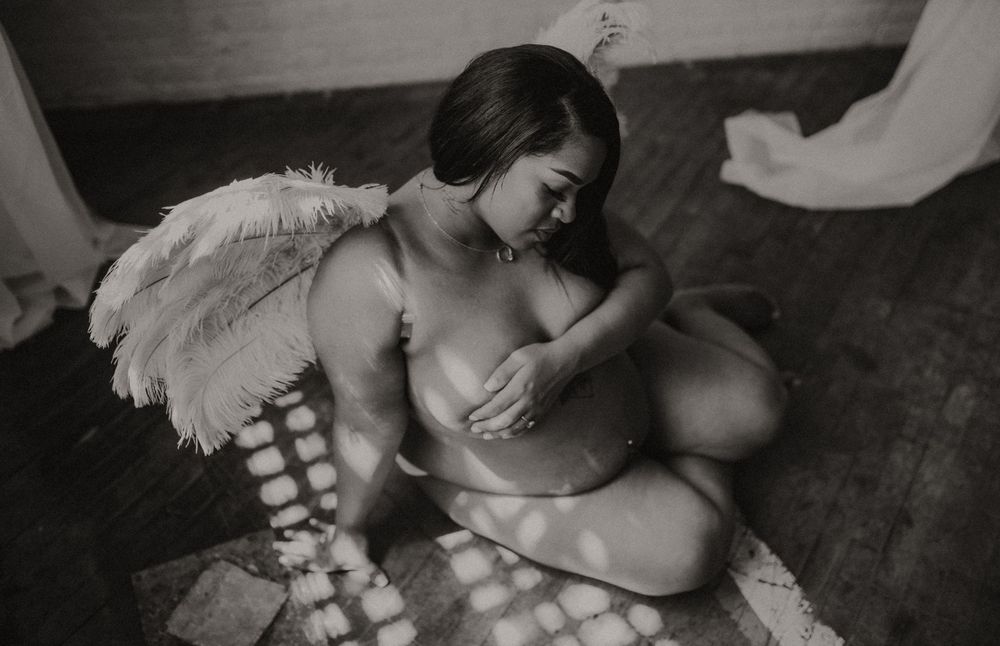Black and white photo of pregnant woman with angel wings sitting on floor in soft natural light.