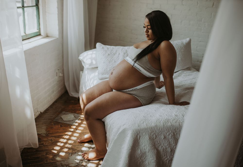 Pregnant woman in grey lingerie sitting on white bed in sunlit bedroom.