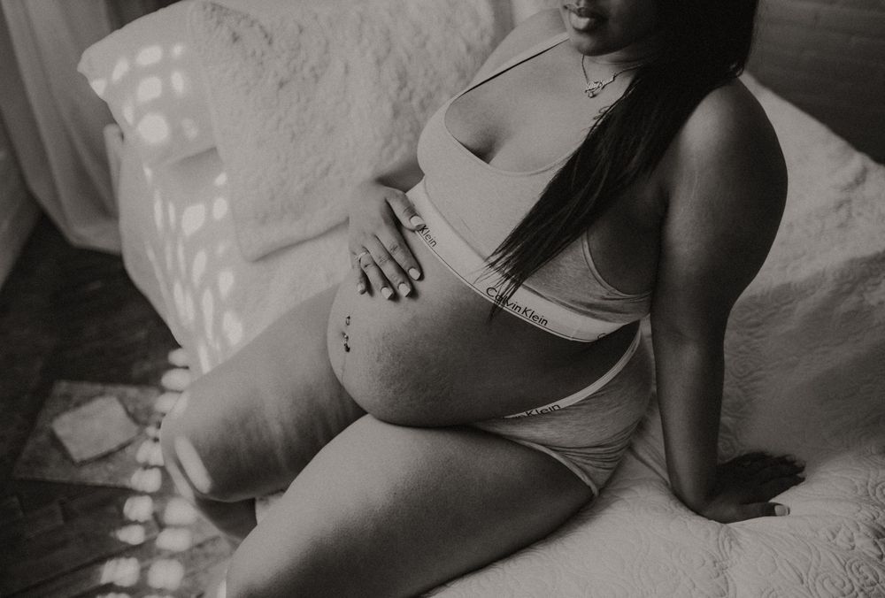 Black and white maternity photo of pregnant woman in lingerie sitting on bed cradling baby bump.
