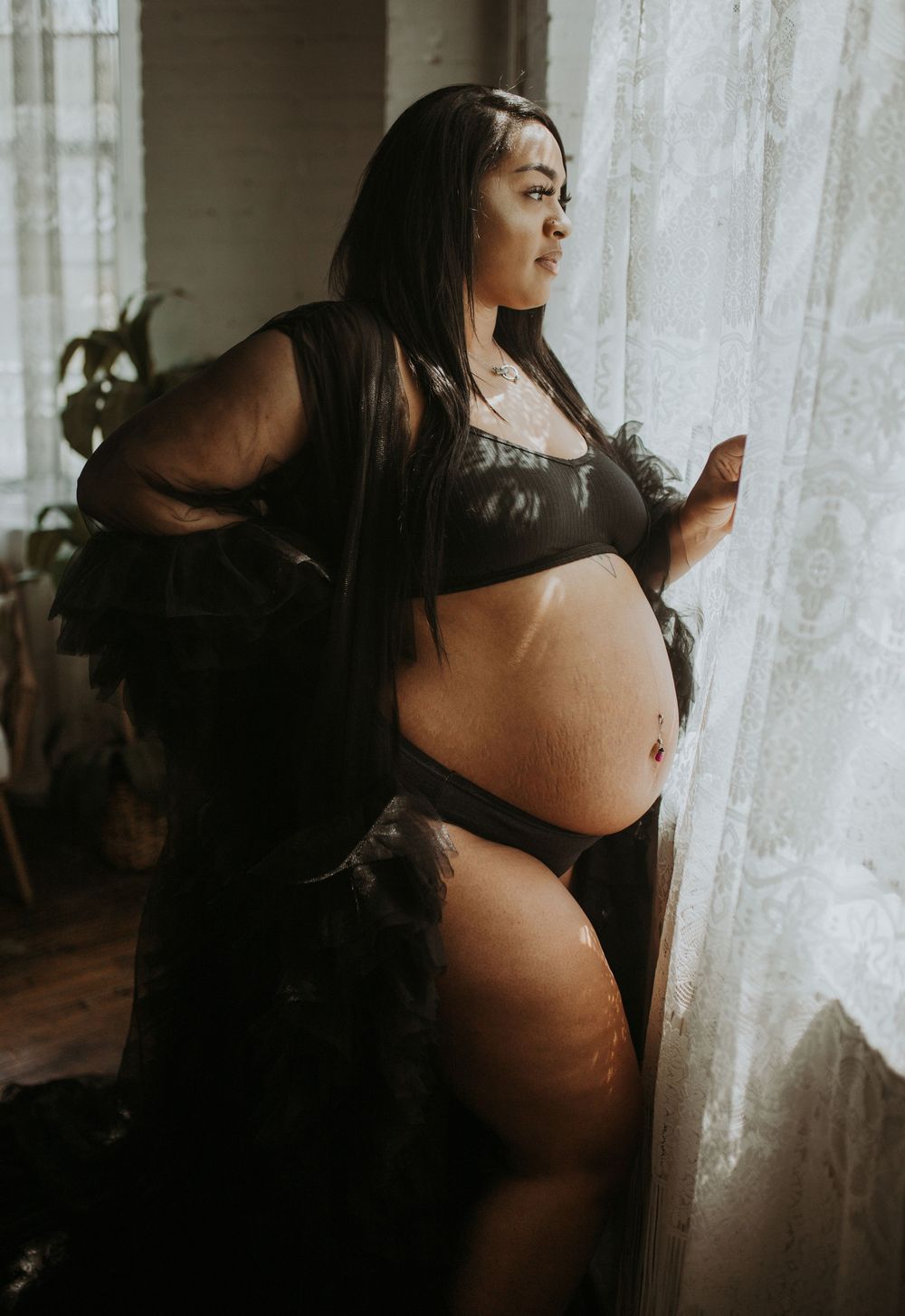 Pregnant woman in black lingerie and sheer robe gazing out window, elegant maternity photo.