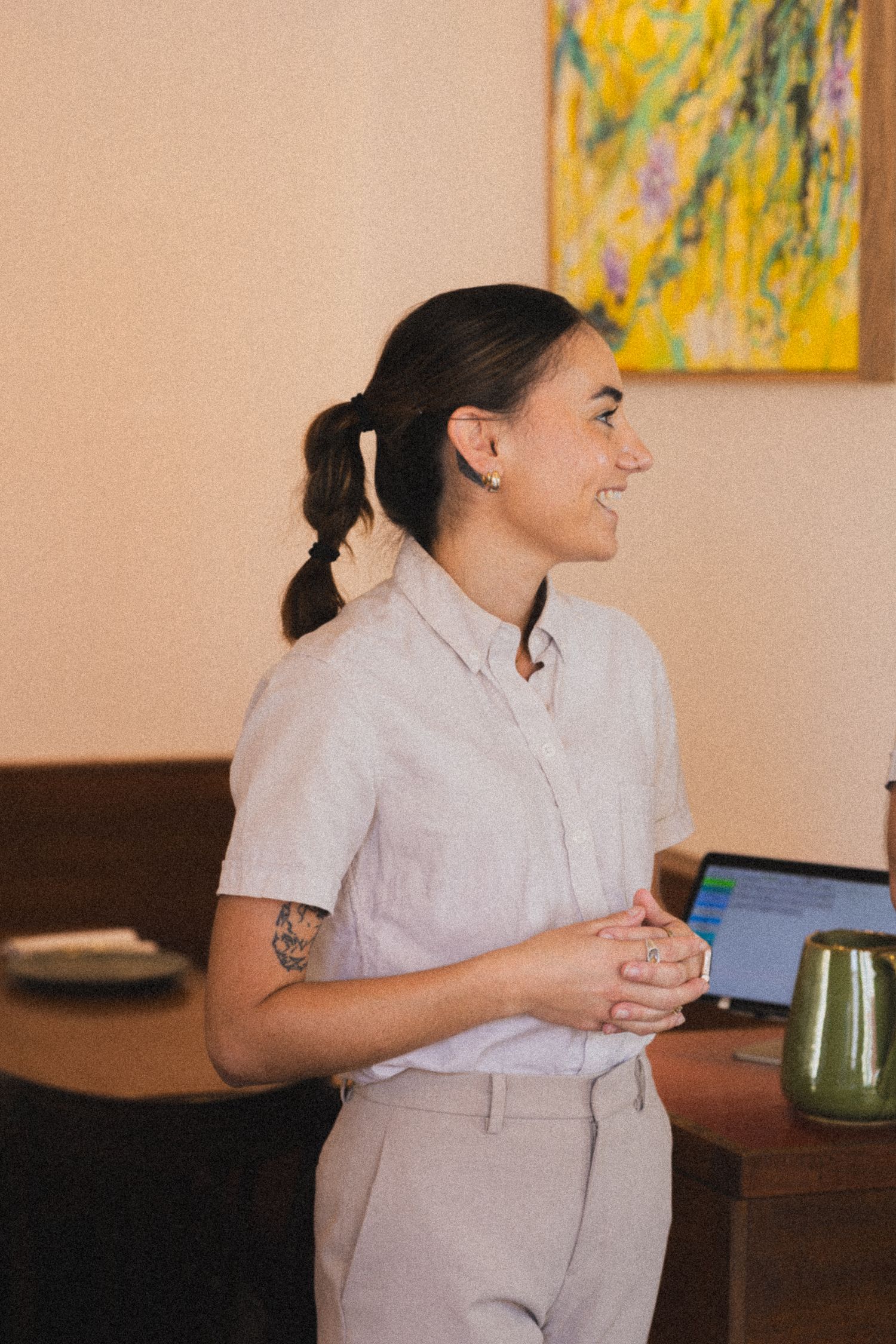 Smiling woman with ponytail in light button-up shirt stands confidently in a warmly lit room with colorful artwork.