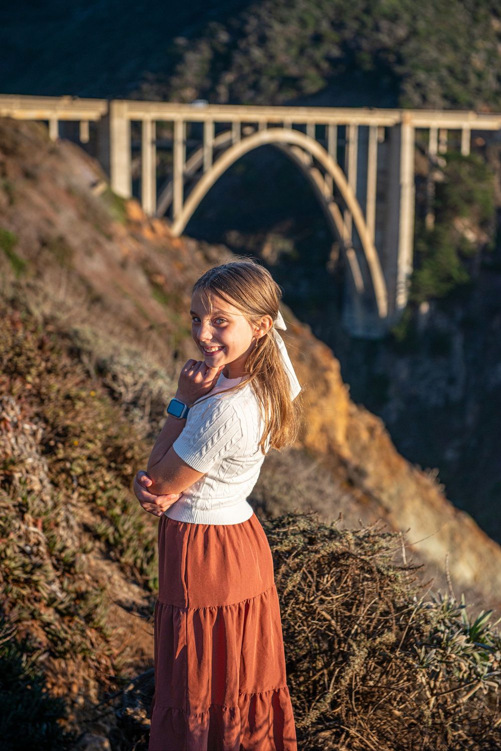 Young girl in white top and rust skirt smiling at Bixby Creek Bridge, Big Sur, California.