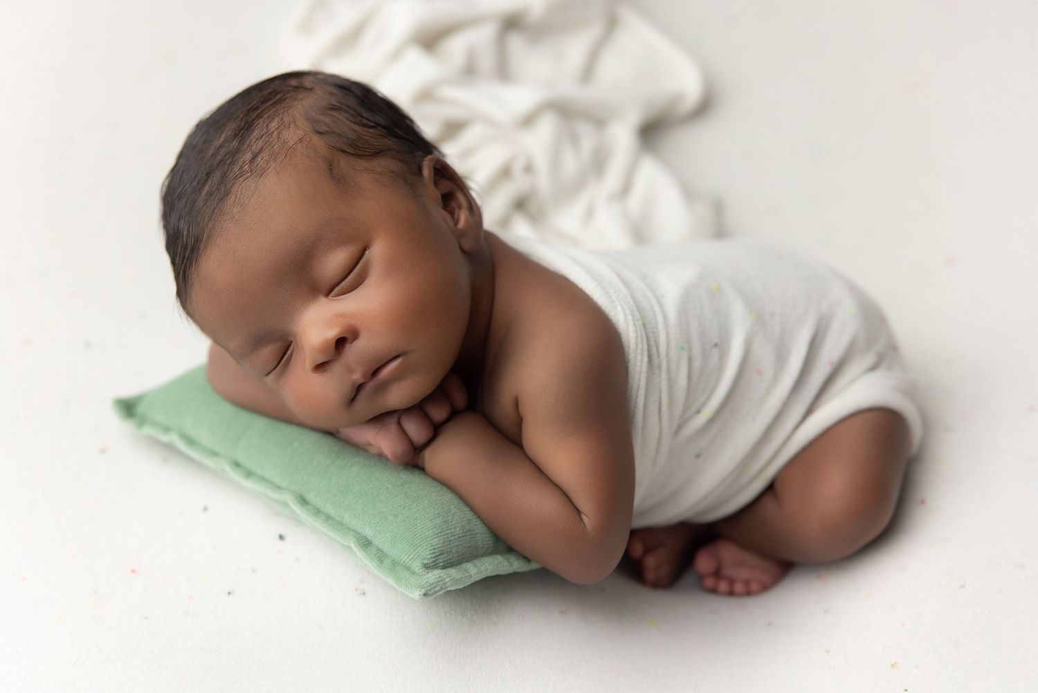 Peaceful newborn baby sleeping on a small green pillow, wrapped in white cloth on a white background.
