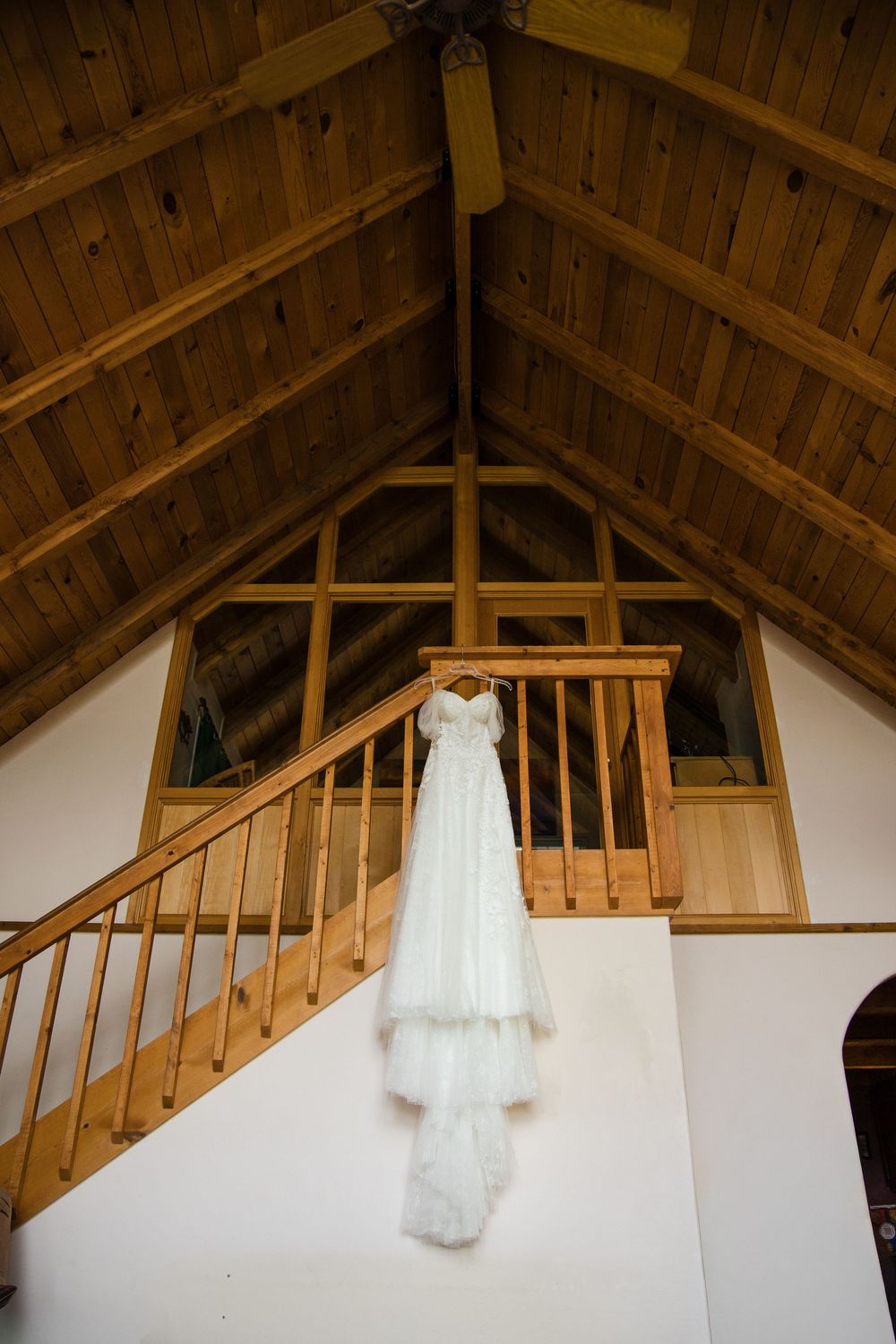 White wedding dress hanging from wooden staircase railing in rustic barn venue with vaulted wood ceiling.