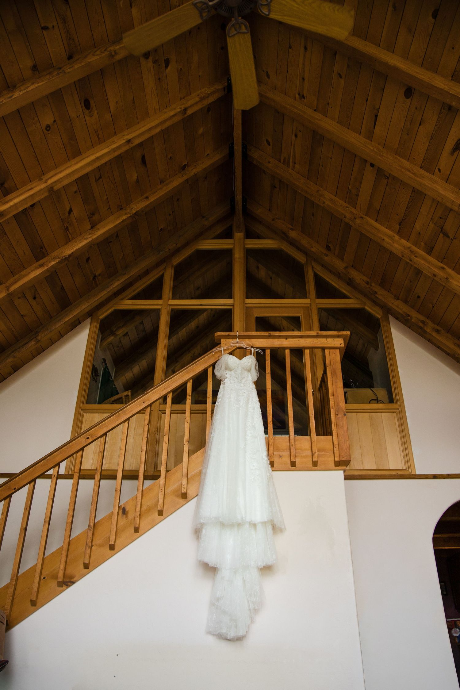 White wedding dress hanging from wooden staircase railing in rustic barn venue with vaulted wood ceiling.