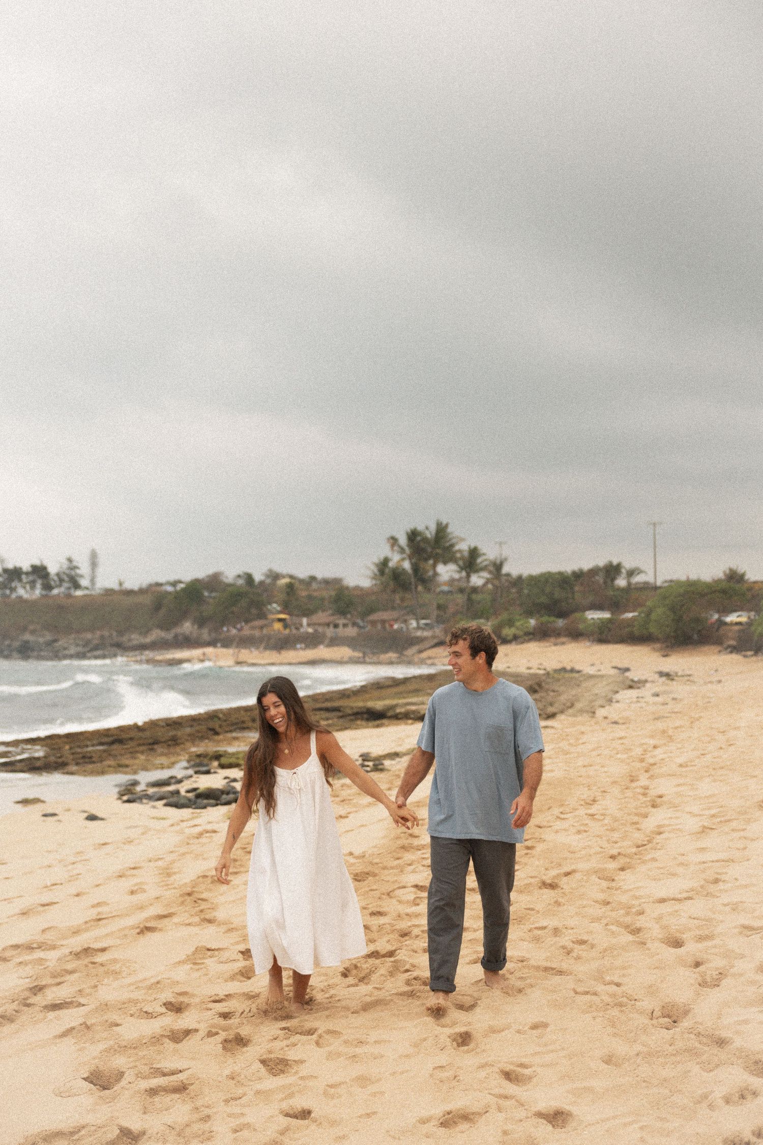 Couple holding hands and walking on sandy beach, woman in white dress and man in blue shirt, overcast sky.