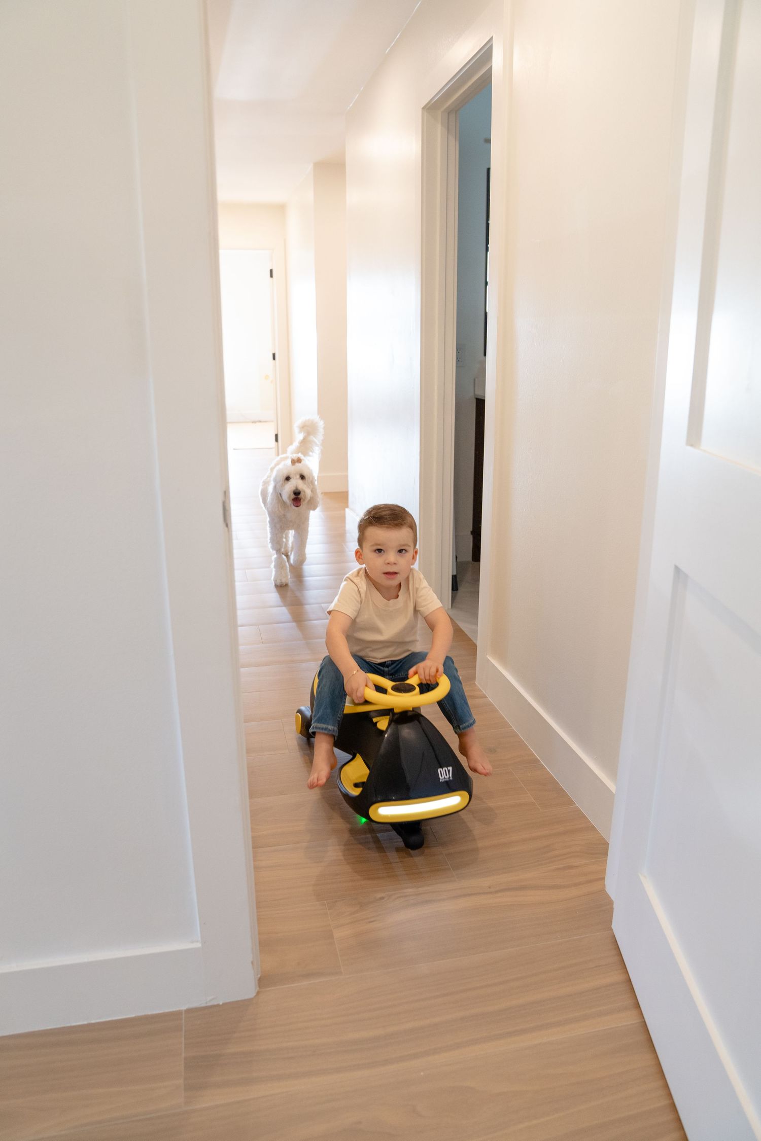 Toddler riding yellow toy car down hallway with white fluffy dog following behind on light wood floors.