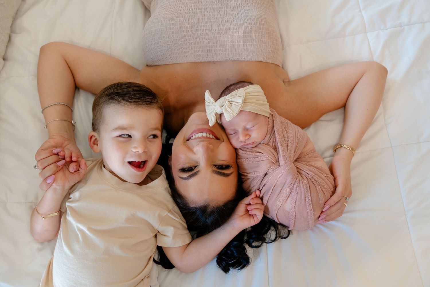 A mother lies on a white bed with her toddler and newborn baby, sharing a tender family moment.