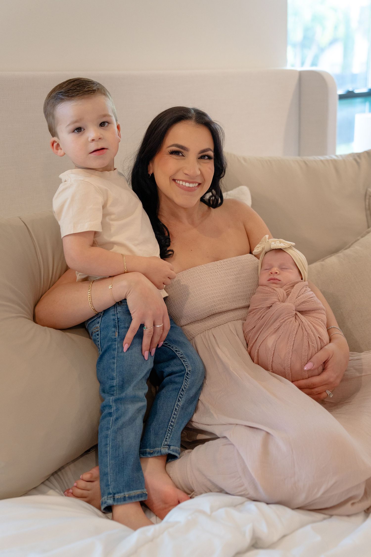 A smiling brunette woman sits on a beige sofa holding a newborn in pink wrap beside a young toddler boy in jeans.