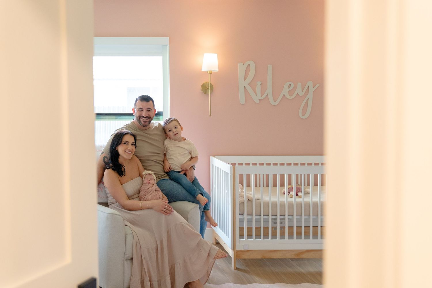 Family of four poses in a pink nursery with a white crib and Riley name sign on the wall.