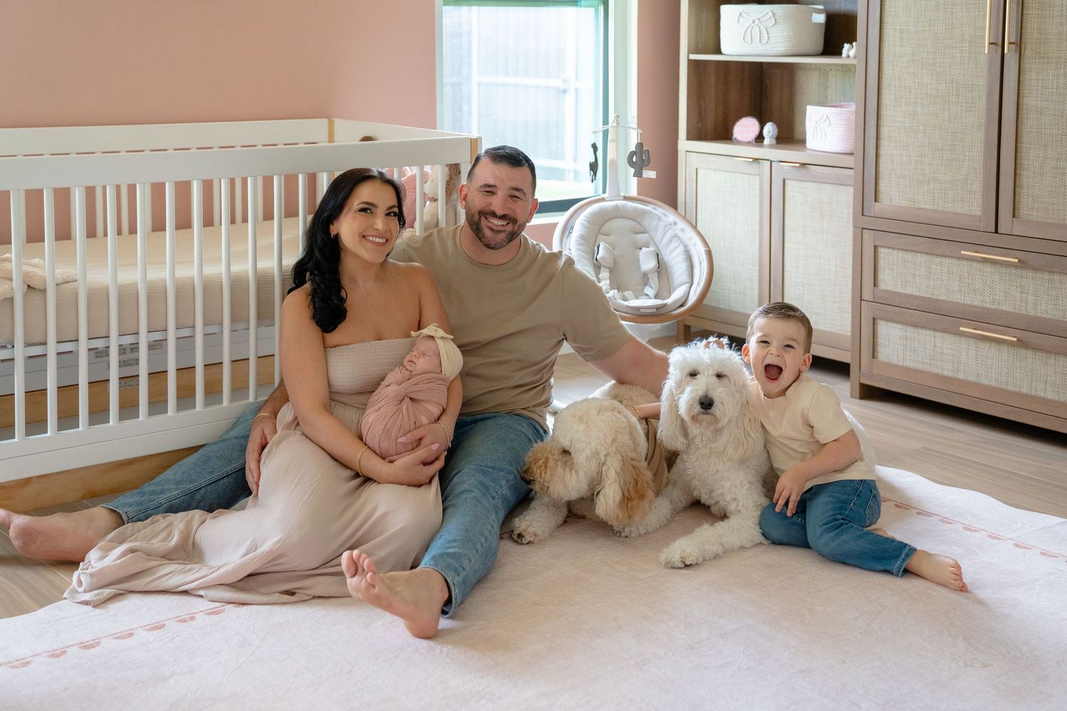 Happy family with pregnant mom, dad, toddler, and fluffy dog sitting together in a cozy nursery room.