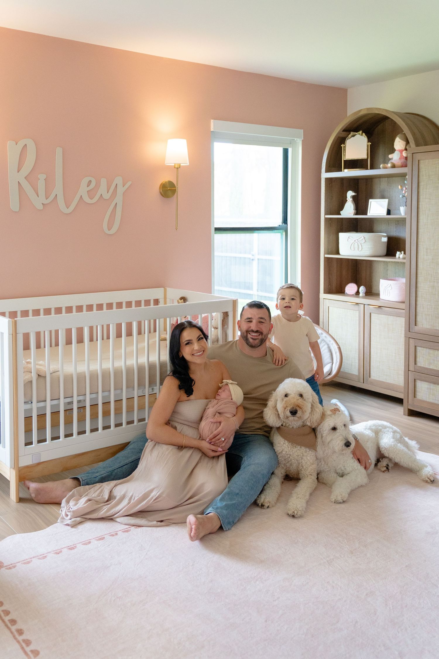 Family with newborn, toddler, and white dog sitting in pink nursery named Riley with white crib and arched bookshelf.