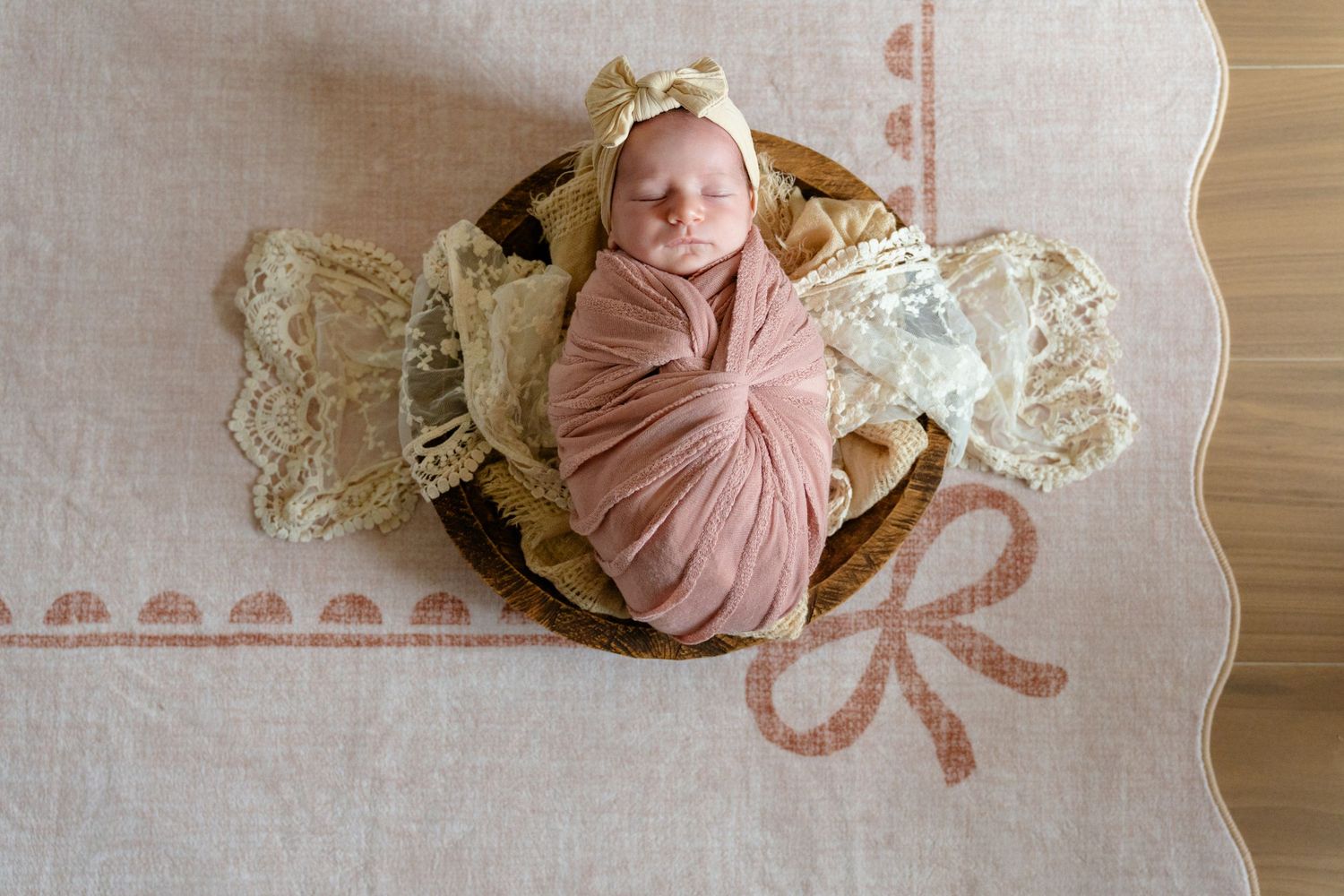 Newborn baby swaddled in pink wrap with bow headband, nestled in wooden bowl on lace and floral rug.
