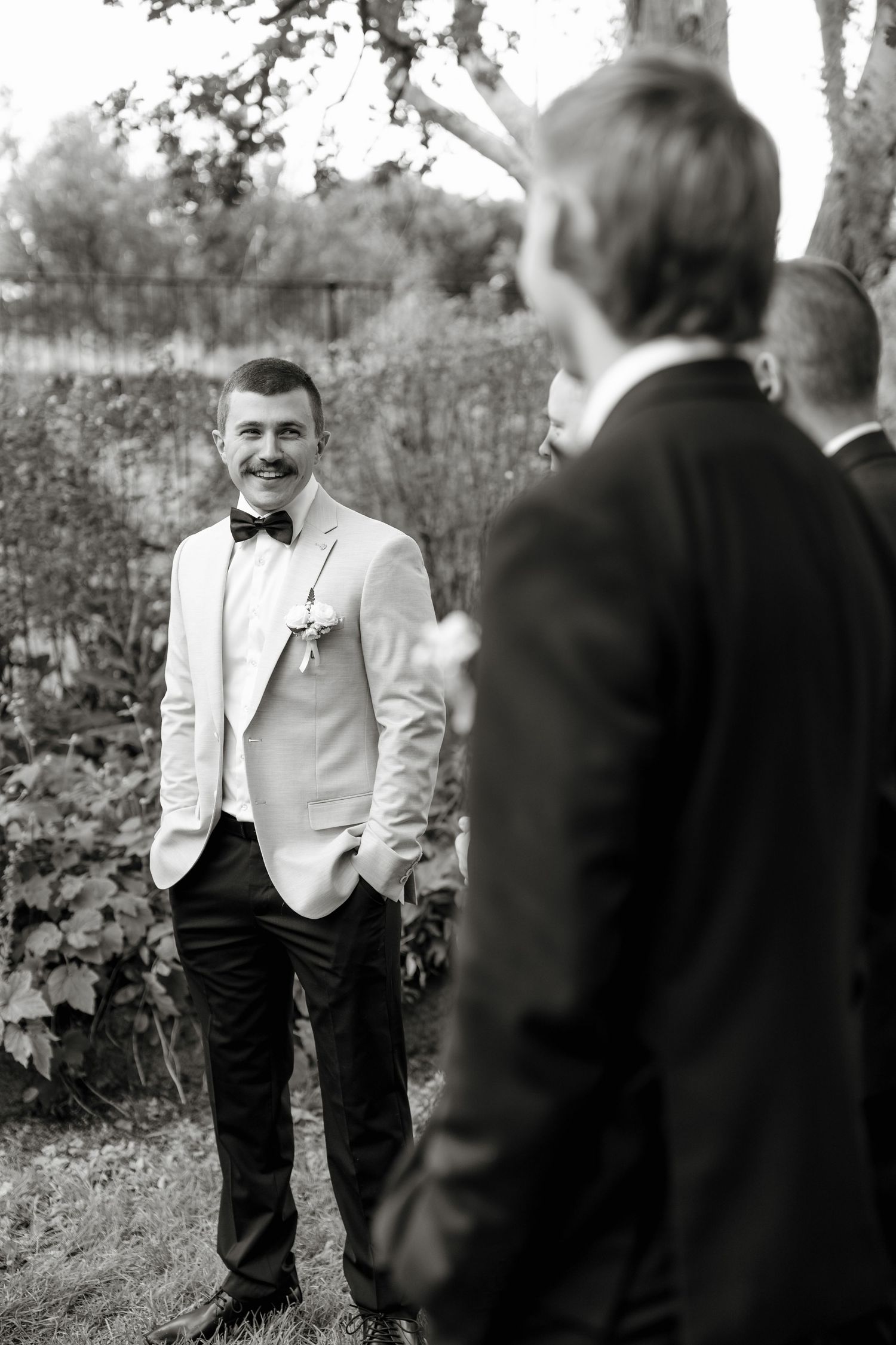 Black and white photo of smiling groom in white tuxedo jacket and bow tie chatting with groomsmen outdoors.