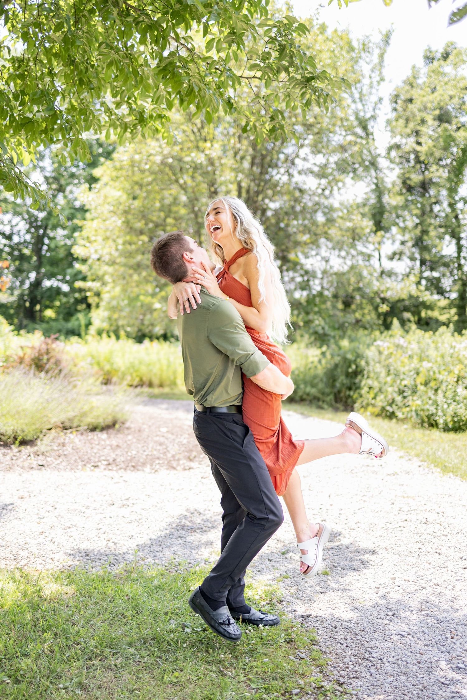 A man lifts a laughing blonde woman in an orange dress on a sunny garden path surrounded by green trees at Voice of America Park.