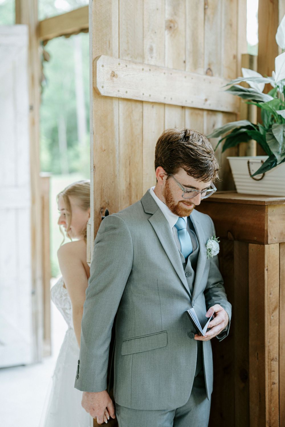 Groom in gray suit reads vows while bride in white wedding dress stands behind wooden barn door during first look.