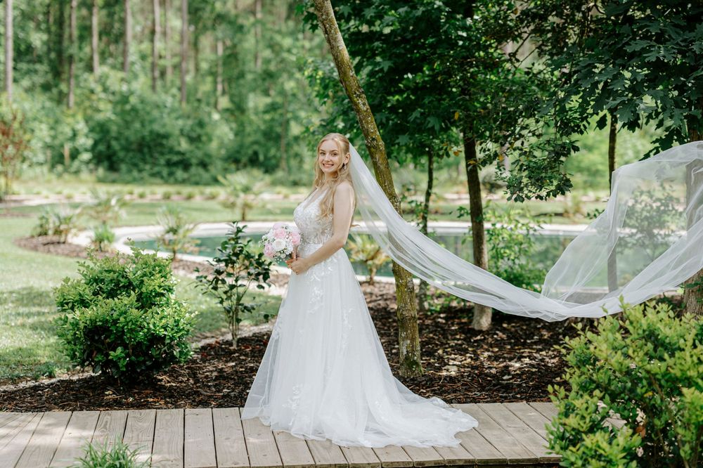 Bride in white gown and flowing veil stands on wooden deck surrounded by lush garden greenery on her wedding day.