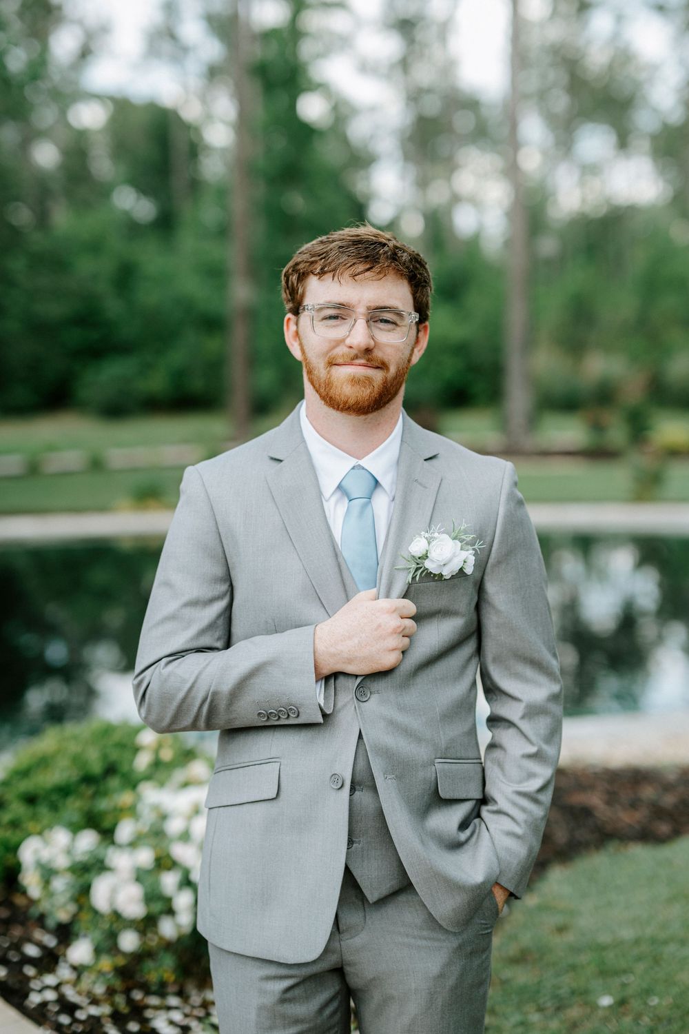 Groom in gray suit with blue tie and boutonniere smiling outdoors at wedding venue.