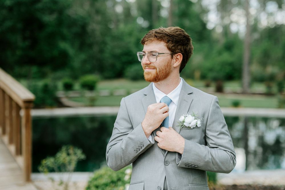 Groom in gray suit adjusting blue tie with boutonniere outdoors at elegant garden wedding venue.