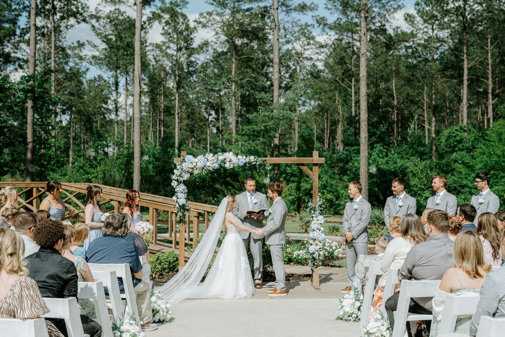 Outdoor wedding ceremony in a forest setting with bride and groom at floral arch, groomsmen in gray suits, and guests seated.