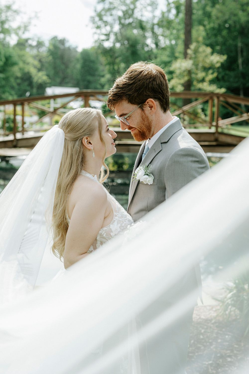 Bride and groom share a romantic moment near a wooden bridge, her veil flowing in the breeze on their wedding day.