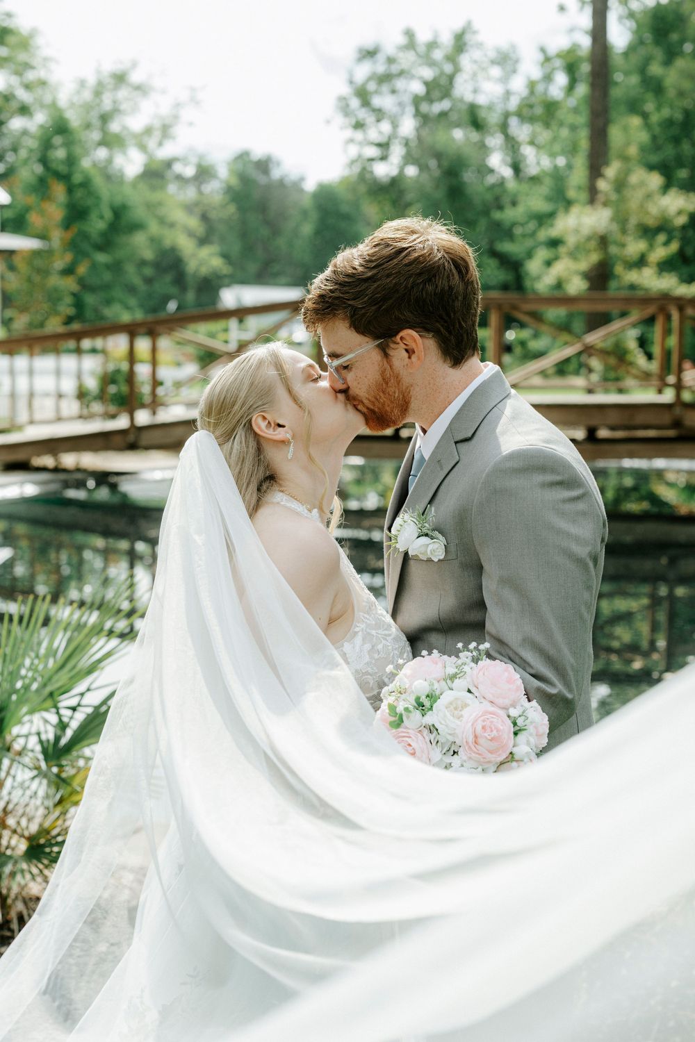 Bride and groom share a romantic kiss outdoors, with bridal veil flowing and pink rose bouquet near a wooden bridge.