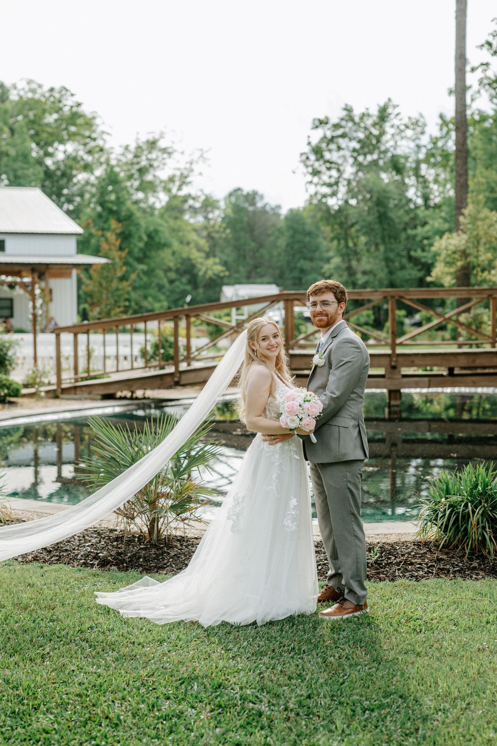 Bride and groom pose together outdoors near a wooden bridge and pond, bride holding pink bouquet with flowing veil.