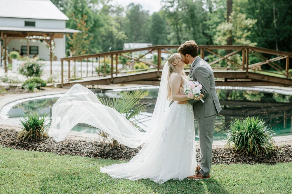 Bride and groom kissing outdoors with flowing veil, rustic pond and wooden bridge backdrop on wedding day.