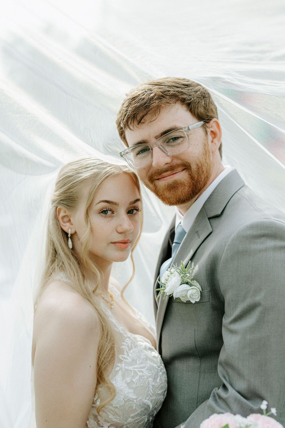 A bride and groom pose romantically under a flowing white veil, groom wearing gray suit with floral boutonniere.