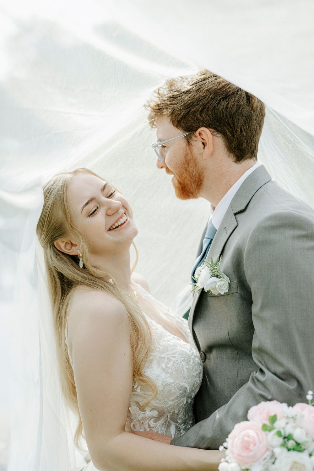 Joyful bride laughs with groom under flowing veil, holding pink roses, both dressed in wedding attire outdoors.