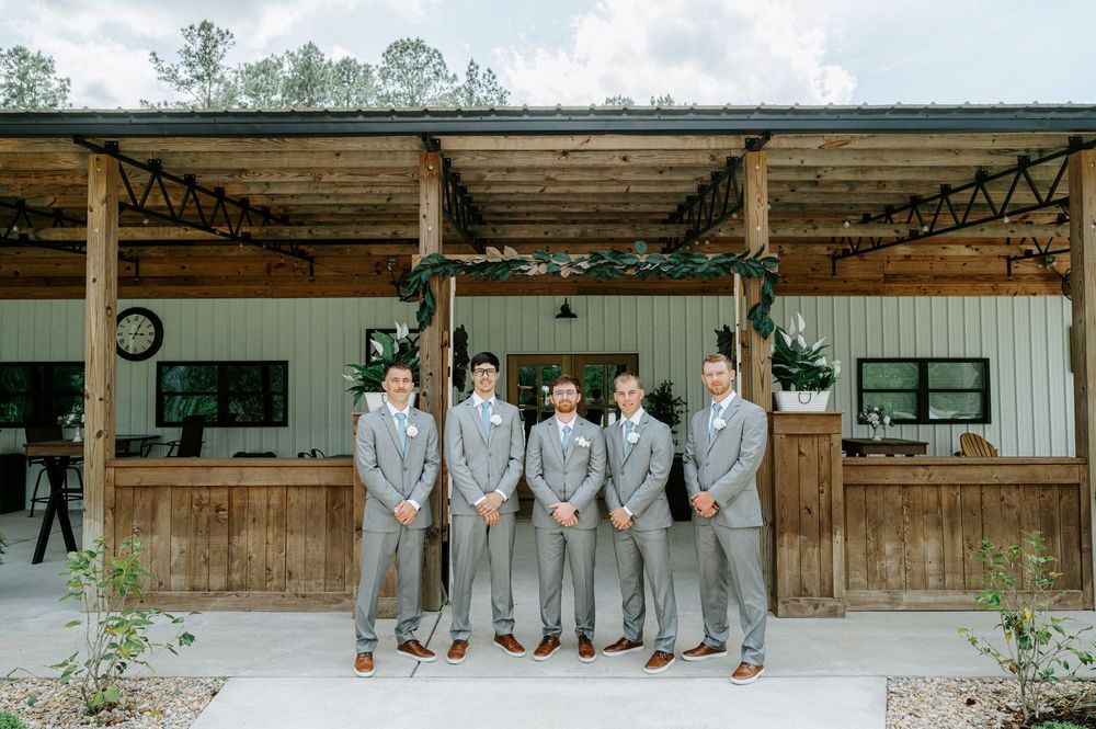 Five groomsmen in matching gray suits with boutonnieres pose in front of a rustic barn wedding venue.