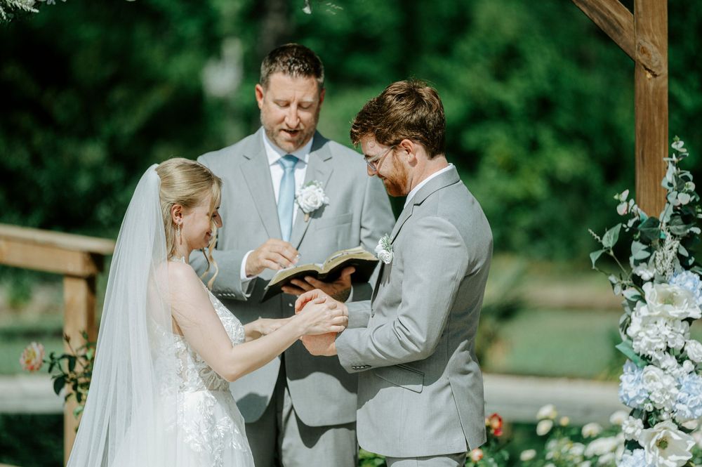 Bride and groom exchange vows outdoors with officiant, surrounded by floral arch during elegant garden wedding ceremony.