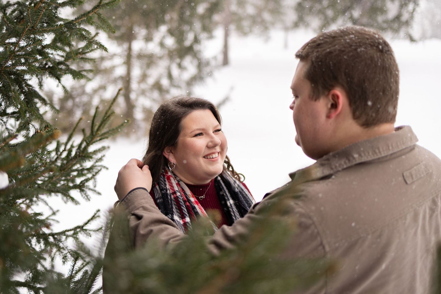 Couple smiling at each other outdoors in snowy winter forest near evergreen trees, romantic engagement photo session.